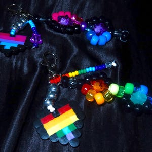 May include: Two beaded keychains featuring pixelated heart charms. One heart has horizontal stripes of pink, purple, and blue, while the other has rainbow stripes. Both include silver-toned lobster clasps and various colorful plastic pony beads.