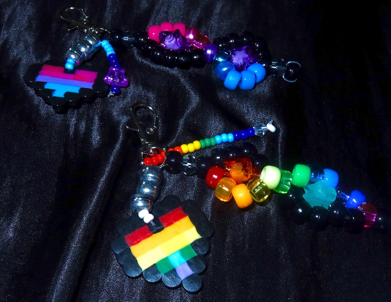 May include: Two beaded keychains featuring pixelated heart charms. One heart has horizontal stripes of pink, purple, and blue, while the other has rainbow stripes. Both include silver-toned lobster clasps and various colorful plastic pony beads.