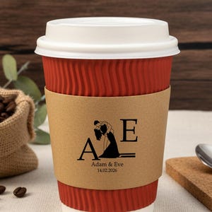 May include: A red paper coffee cup with a white lid and a brown paper sleeve. The sleeve features a black graphic of a couple and the initials "A" and "E" with the date "14.02.2026". Coffee beans and a burlap sack are in the background.