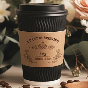 May include: A black paper coffee cup with a decorative sleeve that reads "A Baby is Brewing" with a coffee bean graphic. The cup is surrounded by coffee beans, a wooden spoon, and a floral arrangement.
