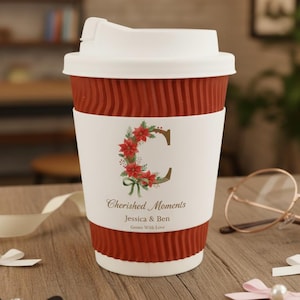 Pode incluir: Uma caneca de café de viagem vermelha e branca com tampa branca. A caneca tem uma faixa branca com um design floral "C" e o texto "Cherished Moments" e nomes. A caneca está sobre uma superfície de madeira.