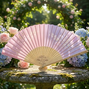 May include: A pink hand fan with a light wood handle. The fan features a floral design and the text "Mia & Liam 18.05.2026". The fan is displayed in front of a backdrop of roses and hydrangeas.