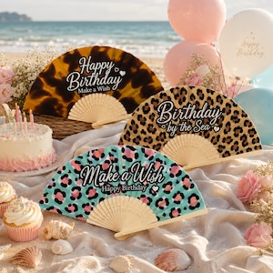 May include: Three folding hand fans with wooden ribs. One has a brown tortoiseshell pattern with "Happy Birthday Make a Wish" text. Another has a leopard print with "Birthday by the Sea" text. The third has a teal and pink leopard print with "Make a Wish Happy Birthday" text.