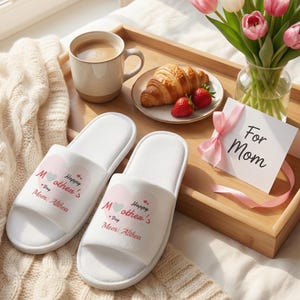以下が含まれることがあります： ピンクのハートのデザインと「Happy Mother's Day」と「Mom: Althea」の文字が入った白い母の日スリッパ。木製のトレイには、コーヒーカップ、クロワッサン、イチゴ、チューリップの花瓶が置かれています。