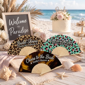 Puede incluir: Tres abanicos plegables con varillas de madera. Uno presenta un estampado de leopardo marrón con el texto "Bridal Shower Future Mrs. Anderson". Otro tiene un estampado de leopardo turquesa y rosa con "Love is in the Air Emily's Bridal Shower". Uno es marrón oscuro con "Cheers to the Bride Emily's Shower".