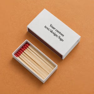 May include: A rectangular white matchbox with a sliding tray containing wooden matches with red tips. The top of the box features the black printed text: Your custom text/design/logo. The box is shown both closed and open to reveal the matches inside.