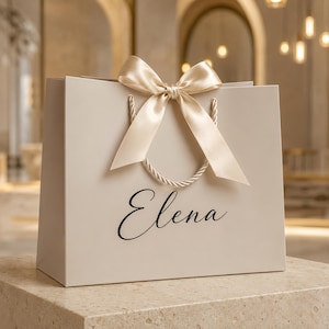 May include: A beige gift bag featuring the name Elena printed in black script lettering. The bag includes twisted rope handles and a large, centered satin ribbon bow in a matching beige color, positioned at the top between the handles.