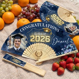 May include: Three graduation fans with a navy blue floral design and the text "Congratulations Class of 2026 Daniel". One fan is open, two are closed. The fans have a photo of a graduate and a wooden handle.