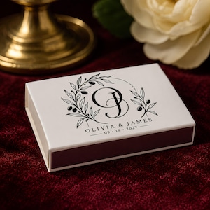 May include: A rectangular white matchbox featuring a black printed design of an olive branch wreath surrounding the intertwined initials OJ. Below the wreath, the text reads OLIVIA & JAMES and the date 09 18 2027 in a clean serif font.