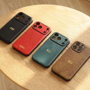 May include: Four leather phone cases in black, red, teal, and brown, each with a gold monogram. The cases are on a light wood surface. The cases have cutouts for the camera and buttons.