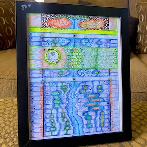 May include: Abstract art piece in a black frame. The artwork features colorful, geometric shapes and patterns in blue, green, orange, and yellow. The piece has a unique, hand-drawn aesthetic, with a variety of lines and symbols.