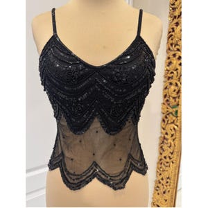 May include: A black sleeveless camisole top featuring a V-neckline and thin spaghetti straps. The bodice is heavily embellished with black sequins and intricate beadwork in floral patterns, transitioning into a sheer mesh hem with a scalloped edge.