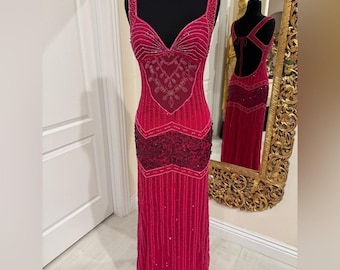 May include: A vibrant red formal gown with a V-neckline and beaded embellishments. The dress features a fitted bodice with intricate beadwork and a flowing skirt. The back of the dress is open with a tie closure. The dress is displayed on a black mannequin.