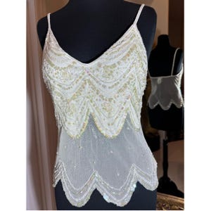 May include: White sequined camisole top with a V-neck and spaghetti straps. The top features a scalloped hem and is embellished with iridescent sequins. The bottom portion is sheer, creating a layered effect. The top is displayed on a black mannequin.