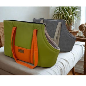 May include: Two pet carrier bags. One is olive green with orange handles and a front pocket. The other is gray with beige handles. Both have a zippered top and are designed for pet transport.