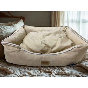 May include: A beige rectangular pet bed with a soft, cushioned interior. The bed has a raised edge with a light gray trim. A small, rectangular tag with the text "LEDUS PREMIUM" is sewn on the front. The bed is designed for comfort and support.