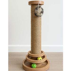 May include: A cat scratching post with a natural rope-wrapped column, topped with a wooden cap and base. The base features three circular platforms with two colorful toy balls. A fluffy, faux fur ball hangs from the top.