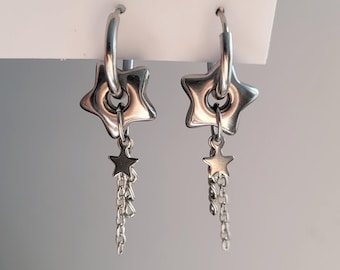 May include: A pair of silver-toned hoop earrings featuring a polished star-shaped charm suspended from each hoop. Attached to the center of each star charm is a smaller star accent connected to a delicate dangling chain, creating a layered effect.