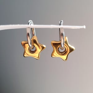 May include: A pair of gold-colored star-shaped earrings with silver hoops. The earrings are hanging against a gray background. The stars have a hole in the center.