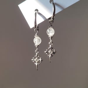 May include: A pair of silver-toned hoop earrings featuring a dangling clear spherical bead and a silver-toned cross-shaped charm with a small embedded crystal at the center. The earrings are attached to a white display card.