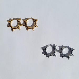 May include: Two pairs of hoop earrings with a spiked design. One pair is gold-colored, and the other is silver-colored. The earrings are displayed on a white background, with the sunlight casting shadows.
