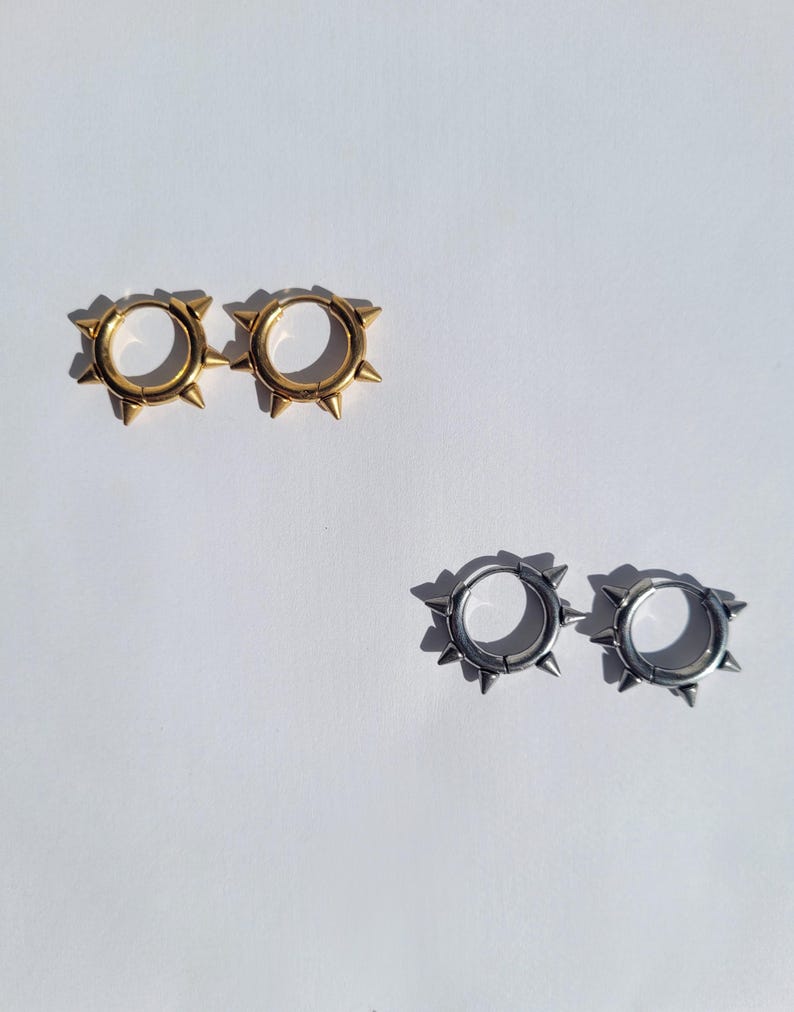 May include: Two pairs of hoop earrings with a spiked design. One pair is gold-colored, and the other is silver-colored. The earrings are displayed on a white background, with the sunlight casting shadows.