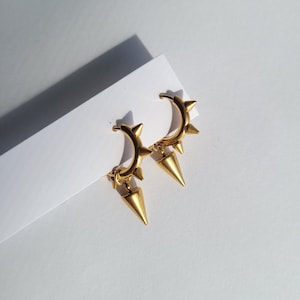 May include: A pair of gold-colored hoop earrings with a spiked design. Each earring features a series of small spikes along the hoop and a larger, dangling spike pendant. The earrings are displayed on a white surface.