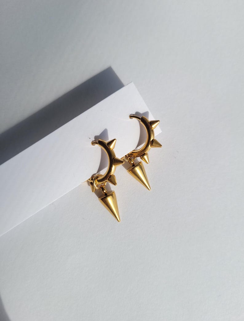 May include: A pair of gold-colored hoop earrings with a spiked design. Each earring features a series of small spikes along the hoop and a larger, dangling spike pendant. The earrings are displayed on a white surface.