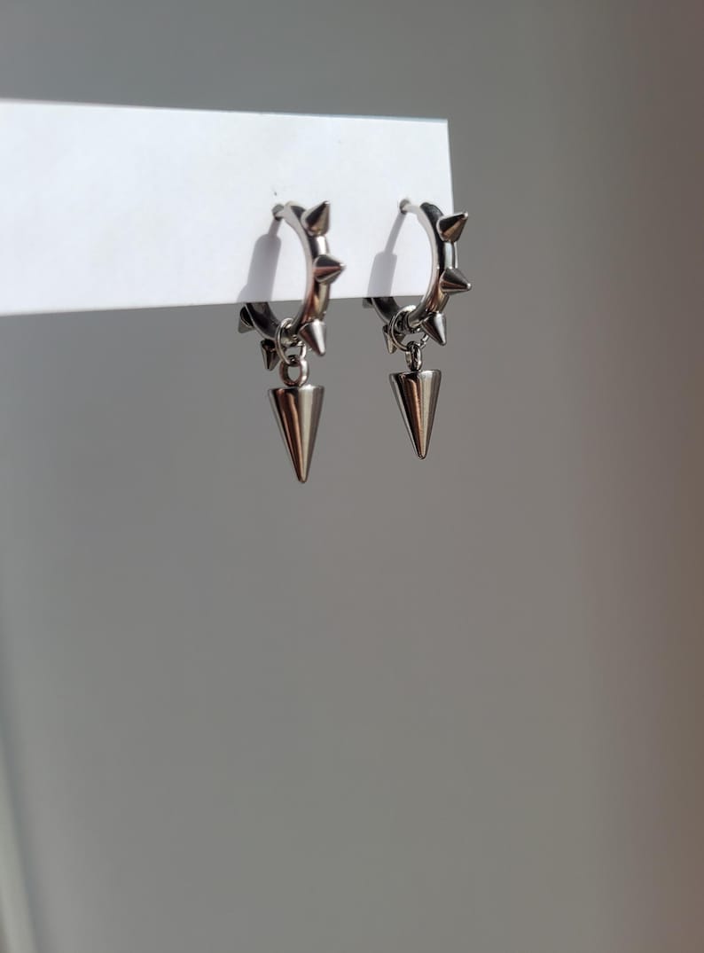May include: A pair of silver-colored hoop earrings. Each earring features small spikes around the hoop and a dangling, cone-shaped pendant. The earrings are displayed against a white background.