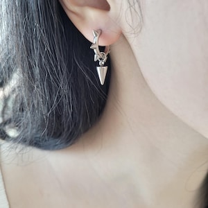 May include: A silver hoop earring with a star-shaped design and a dangling cone-shaped pendant. The earring is worn on the ear, showcasing its edgy and modern aesthetic. The earring's design is detailed and eye-catching.