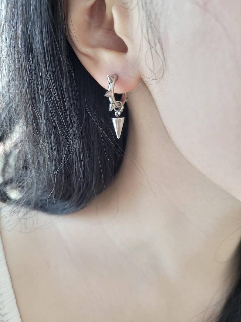 May include: A silver hoop earring with a star-shaped design and a dangling cone-shaped pendant. The earring is worn on the ear, showcasing its edgy and modern aesthetic. The earring's design is detailed and eye-catching.