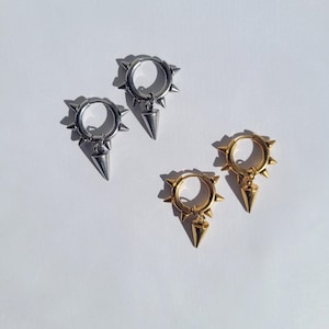 May include: Four earrings, two silver and two gold, are displayed on a white surface. Each earring has a circular hoop design with small spikes around the edge and a dangling cone-shaped charm. The earrings are a punk-inspired jewelry.