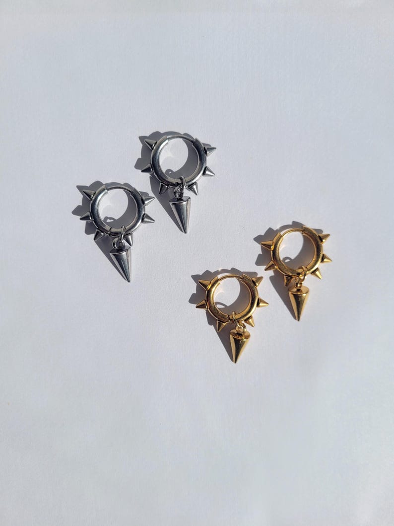 May include: Four earrings, two silver and two gold, are displayed on a white surface. Each earring has a circular hoop design with small spikes around the edge and a dangling cone-shaped charm. The earrings are a punk-inspired jewelry.
