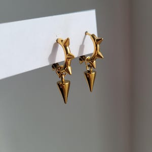 May include: A pair of gold-colored hoop earrings with a spiked design. Each earring features small spikes around the hoop and a dangling, cone-shaped charm. The earrings are displayed against a white background.