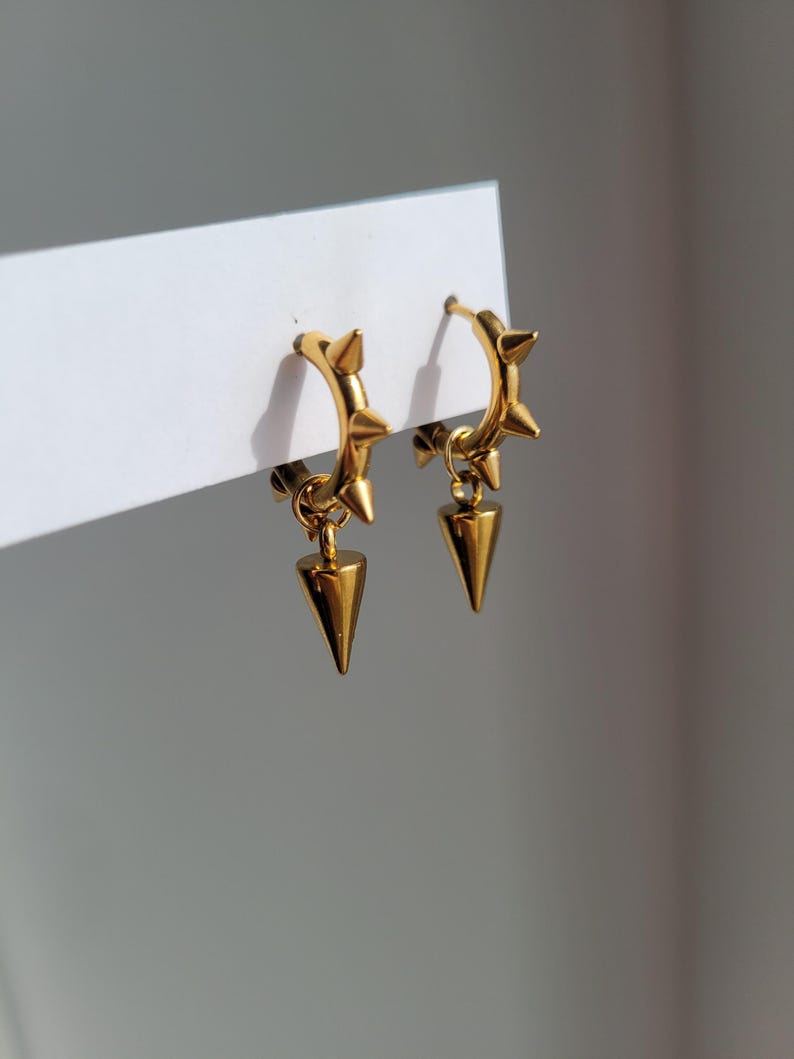 May include: A pair of gold-colored hoop earrings with a spiked design. Each earring features small spikes around the hoop and a dangling, cone-shaped charm. The earrings are displayed against a white background.