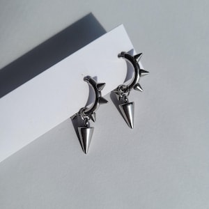 May include: A pair of silver-colored hoop earrings with a punk rock aesthetic. Each earring features a curved hoop adorned with small spikes and a dangling, cone-shaped pendant. The earrings are displayed on a white surface, with a shadow.