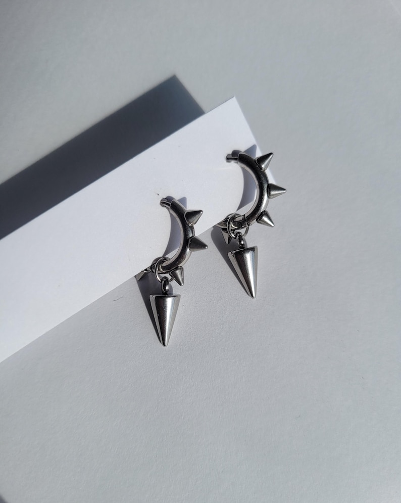 May include: A pair of silver-colored hoop earrings with a punk rock aesthetic. Each earring features a curved hoop adorned with small spikes and a dangling, cone-shaped pendant. The earrings are displayed on a white surface, with a shadow.
