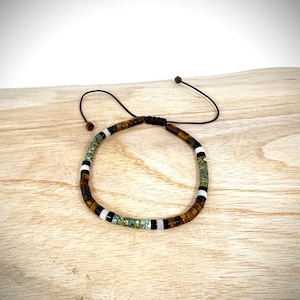 Peut inclure: Bracelet réglable composé de perles marron, noires, blanches et turquoise. Les perles sont enfilées sur un cordon marron foncé, avec une fermeture coulissante. Le bracelet est présenté sur une surface en bois.