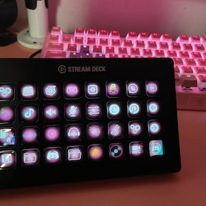 Könnte beinhalten: Schwarzes Stream Deck mit beleuchteten Tasten, die verschiedene Symbole anzeigen, darunter Logos von Social-Media- und Streaming-Apps. Die Tastatur im Hintergrund hat pinkfarbene, hintergrundbeleuchtete Tasten. Das Stream Deck befindet sich auf einer rosa Oberfläche.