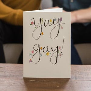 May include: A greeting card with the words "Yay gay!" in elegant script, adorned with floral accents. The card is on a wooden table, with two people in the background. One person is wearing a mustard yellow sweater, and the other is wearing a denim shirt.