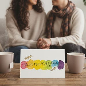 May include: A wooden coffee table holds two taupe mugs with steam rising, and a greeting card that reads "anniversary" in colorful watercolor. Two people are seated on a light-colored sofa in the background.