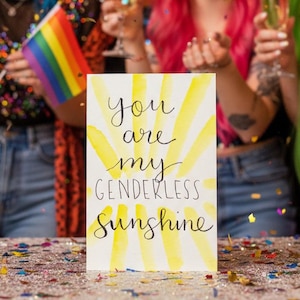 May include: A group of people with brightly colored hair celebrate with champagne and confetti. A sign reads "You are my Genderless Sunshine" in script lettering with a yellow watercolor sunburst design. A rainbow flag is also visible.