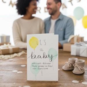 May include: A baby announcement card with the text "it's a baby" and watercolor balloons. The card is on a wooden table with baby booties and gifts. A couple is blurred in the background.