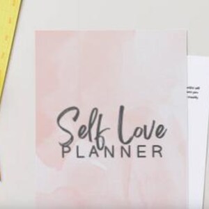 Könnte beinhalten: Ein "Self Love Planner" mit einem rosa Aquarellhintergrund und dem Titel in eleganter Schrift. Ein leuchtend orangefarbener Stift mit einem flauschigen Pomponaufsatz liegt auf einem gelben Notizblock.