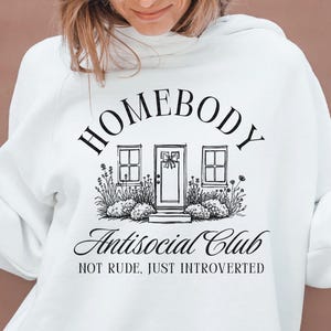 May include: White hoodie featuring the words "HOMEBODY Antisocial Club" in black script. The design includes a house graphic with windows and a door, surrounded by plants. The phrase "NOT RUDE, JUST INTROVERTED" is below the main text.