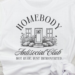 May include: White t-shirt featuring a black illustration of a house with the words "HOMEBODY" and "Antisocial Club". The text "NOT RUDE, JUST INTROVERTED" is also included. The shirt is made of a soft material and is suitable for casual wear.