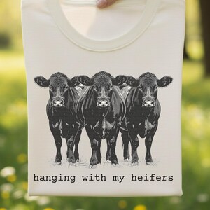 May include: Cream-colored t-shirt featuring a graphic of three black cows. The cows are standing side-by-side, with the text "hanging with my heifers" printed below the image. The shirt has a crew neck.