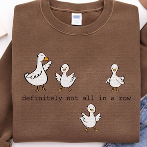 May include: A brown sweatshirt featuring a whimsical design of four white cartoon geese with orange beaks and feet. The text "definitely not all in a row" is printed below the geese. The sweatshirt is made of a soft material.