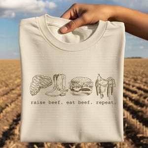 May include: A cream-colored t-shirt featuring a line drawing of a steak, cowboy boot, hamburger, and saddle. The text "raise beef. eat beef. repeat." is printed below the images. The shirt is held up against a background of a harvested field.