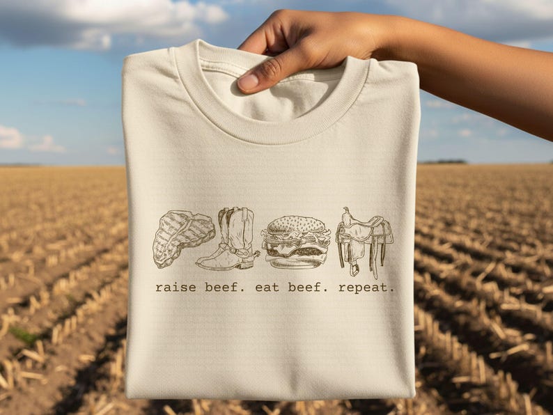 May include: A cream-colored t-shirt featuring a line drawing of a steak, cowboy boot, hamburger, and saddle. The text "raise beef. eat beef. repeat." is printed below the images. The shirt is held up against a background of a harvested field.